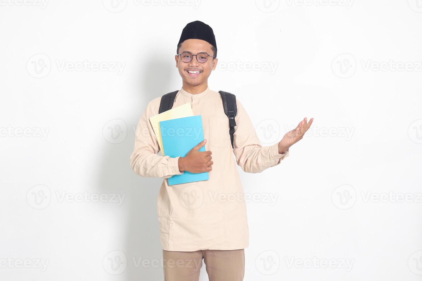 portret van opgewonden leerling Aziatisch moslim Mens in koko overhemd met kalotje draag- rugzak, Holding school- boeken, richten naar de kant. Islamitisch onderwijs concept. geïsoleerd beeld Aan wit achtergrond foto