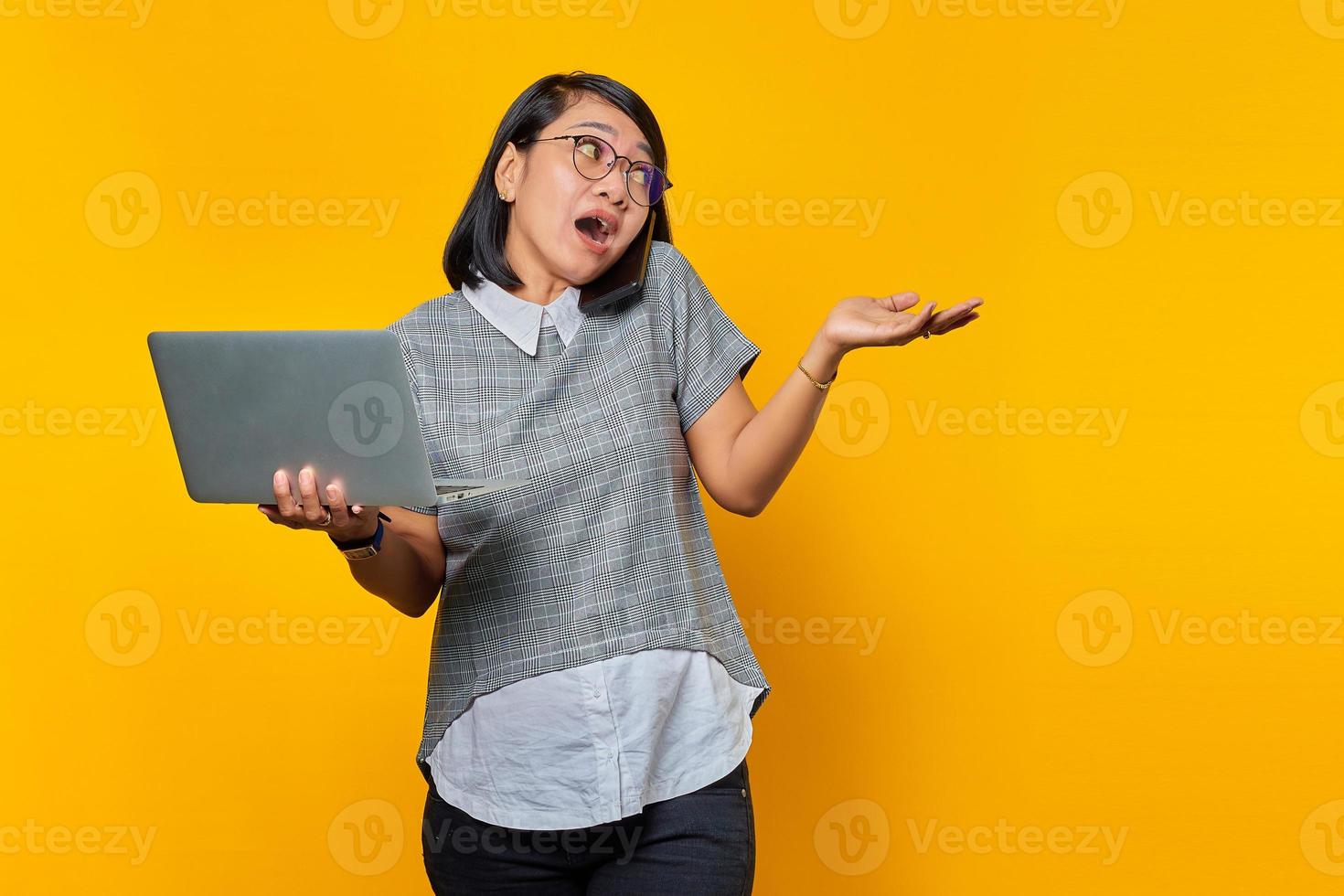 portret van een boze aziatische vrouw die op een smartphone praat terwijl ze een laptop op een gele achtergrond houdt foto
