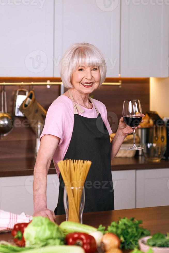 senior vrolijke vrouw drinkt rode wijn tijdens het koken in de moderne keuken. voedsel, onderwijs, levensstijlconcept foto