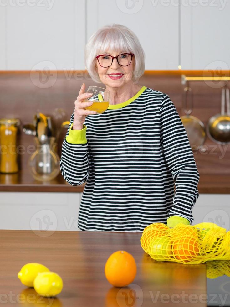 vrolijke, mooie senior lachende vrouw in gestreepte trui die sinaasappelsap drinkt terwijl ze in de keuken staat. gezonde, sappige levensstijl, thuis, senior mensen concept. foto
