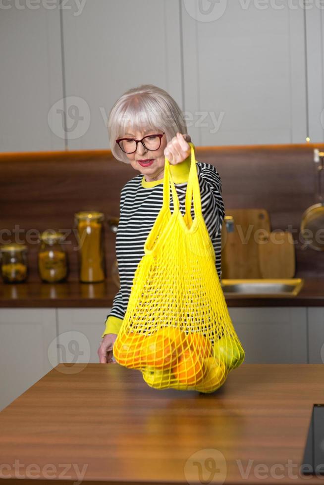 vrolijke mooie senior lachende vrouw in gestreepte trui die citroenen vasthoudt voor limonade terwijl ze in de keuken staat. gezonde, sappige levensstijl, thuis, senior mensen concept. foto