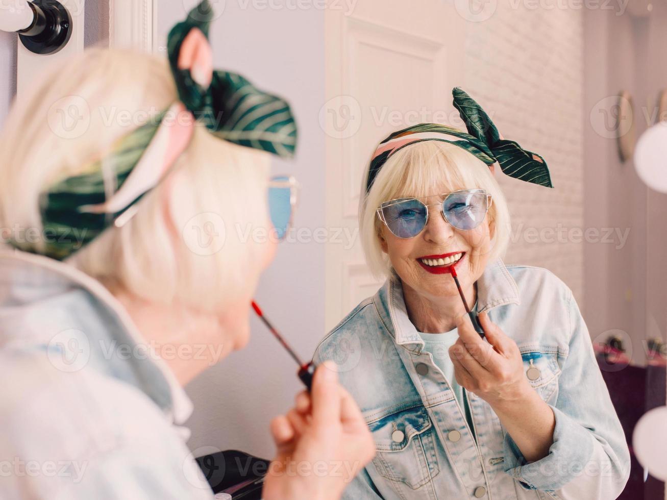 lachende oudere senior stijlvolle vrouw in blauwe zonnebril en jeans jasje met rode lippenstift bij de spiegel in stijlvol loft interieur. stijl, mode, make-up, anti-age concept foto