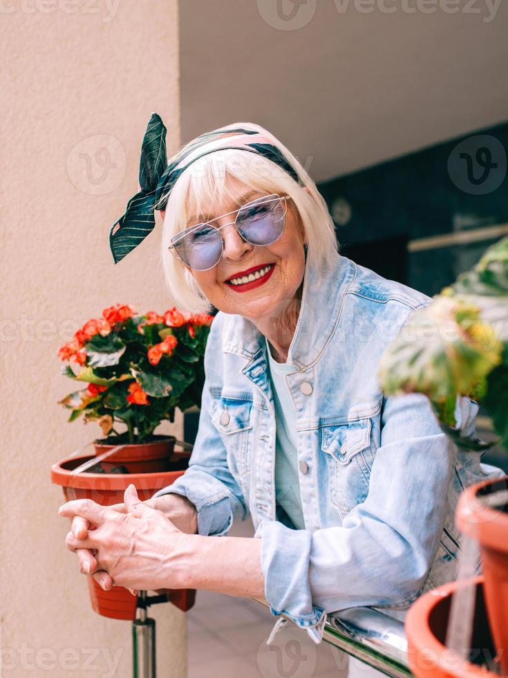 senior stijlvolle vrouw met grijs haar en in blauwe bril en denim staande op balkon buitenshuis. foto