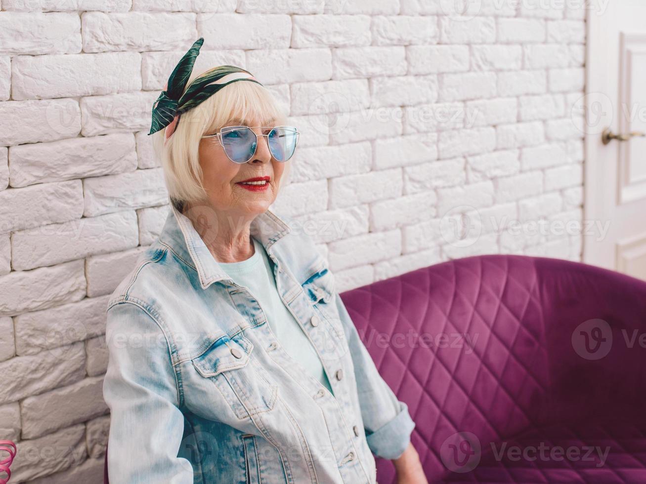 oudere senior stijlvolle vrouw in blauwe zonnebril en spijkerjasje zittend op de bank in stijlvol loft interieur in salon. stijl, mode, wacht, anti-leeftijd concept foto