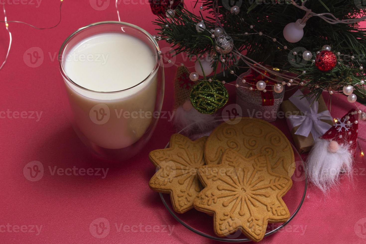 peperkoek en melk voor de kerstman. kerstcompositie met peperkoekkoekjes en melk op een roze achtergrond met een kerstboom en een cadeau. foto