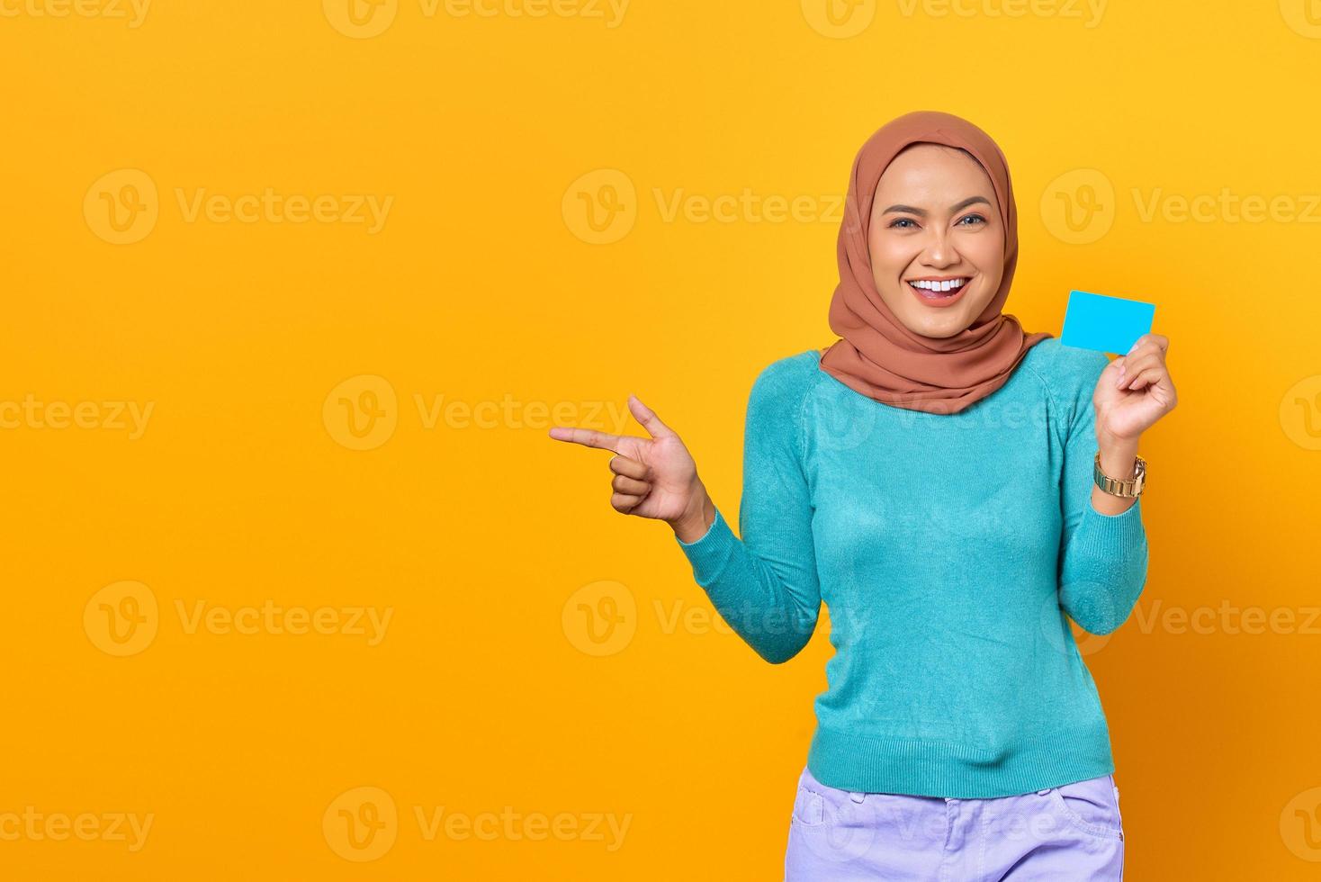 glimlachende aziatische vrouw die een creditcard vasthoudt terwijl ze met de vinger wijst naar kopieerruimte op gele achtergrond foto