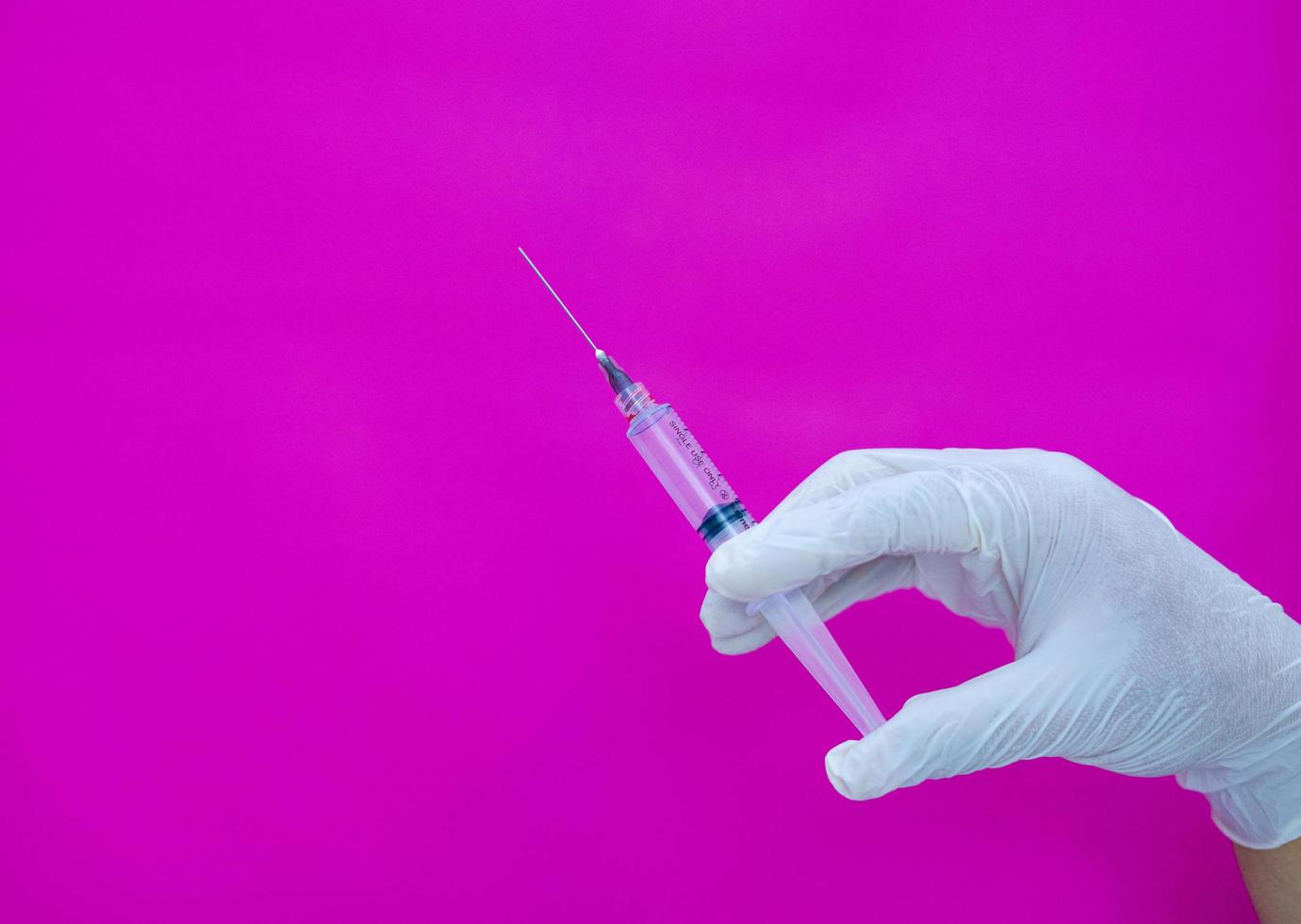 een gehandschoende hand op een roze achtergrond, met een medische spuit die wordt gebruikt om medicijnen en vitamines in het lichaam te injecteren. foto