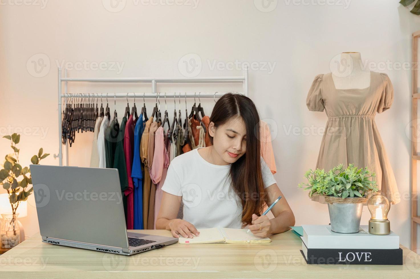 Aziatische vrouw ondernemer van kledingwinkel werken en noteren in notitieboekje met laptop op bureau foto