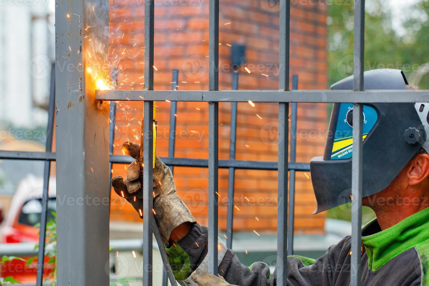 een lasser die een beschermende helm en handschoenen draagt, last een metalen hek rond een flatgebouw. foto