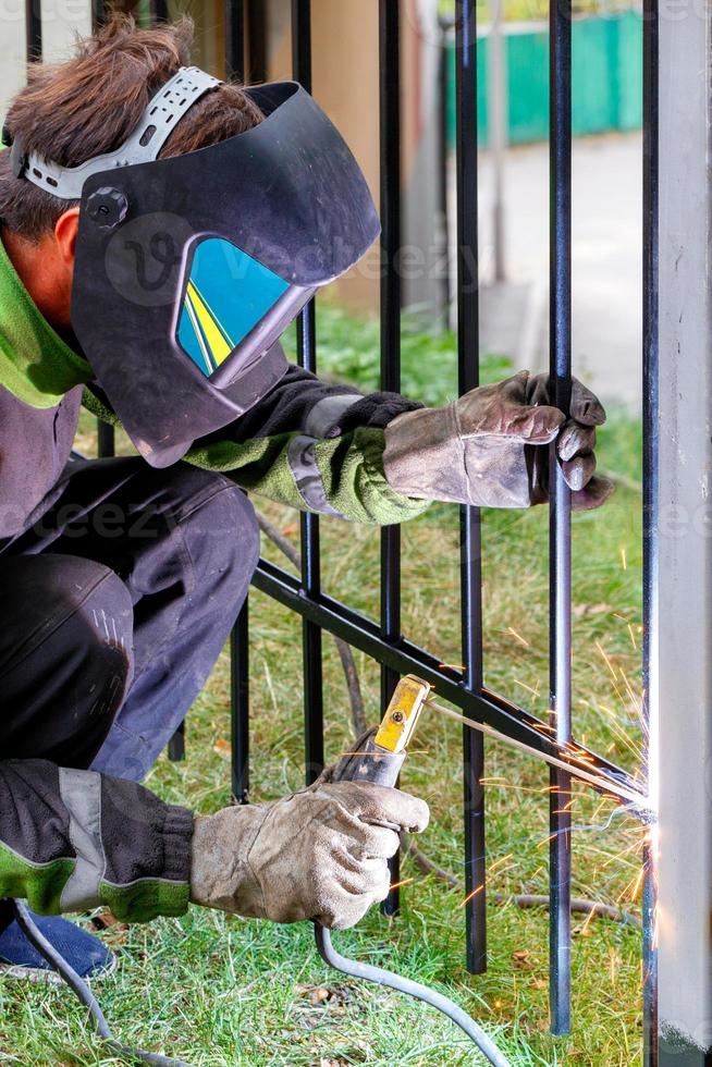 een lasser in een beschermende helm en leren handschoenen is een metalen hek aan het lassen met een elektrode. foto