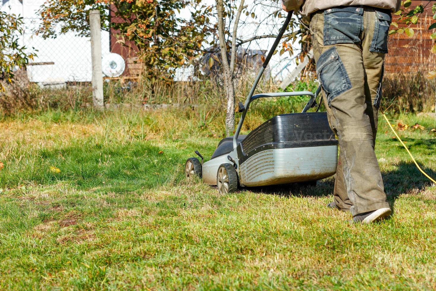 een tuinman maait groen gras met een elektrische grasmaaier in zijn achtertuin op een zonnige herfstdag, achteraanzicht. foto