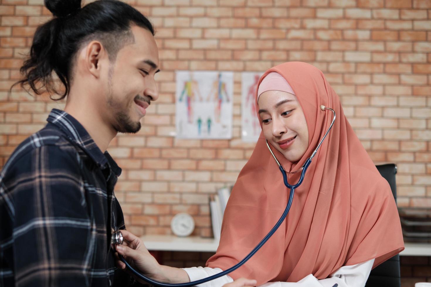 jonge mooie vrouw arts is gezondheid die mannelijke patiënt onderzoekt in het kantoor van de ziekenhuiskliniek en met een glimlach adviseert over medicijnen. deze aziatische medisch specialist is een islamitisch persoon die een hijab draagt. foto