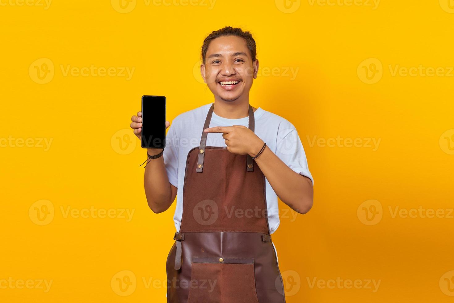 Glimlachende knappe jongeman die naar een leeg smartphonescherm wijst op een gele achtergrond foto