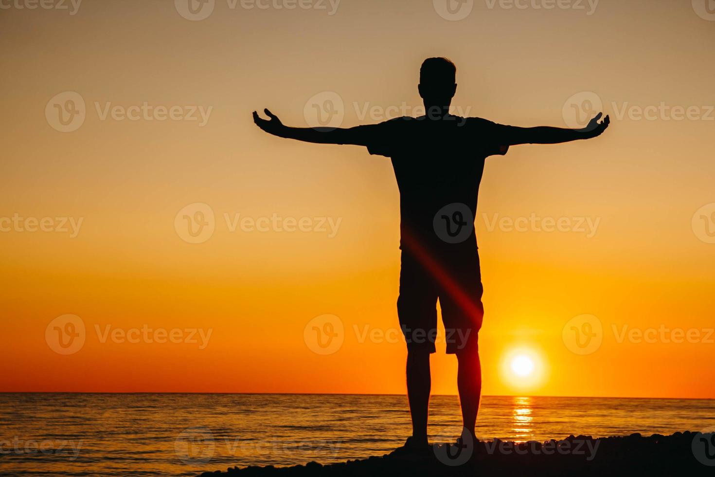 silhouet van een man staat en overweegt op het strand op de achtergrond van de zee en de zonsondergang gooit zijn handen opzij foto