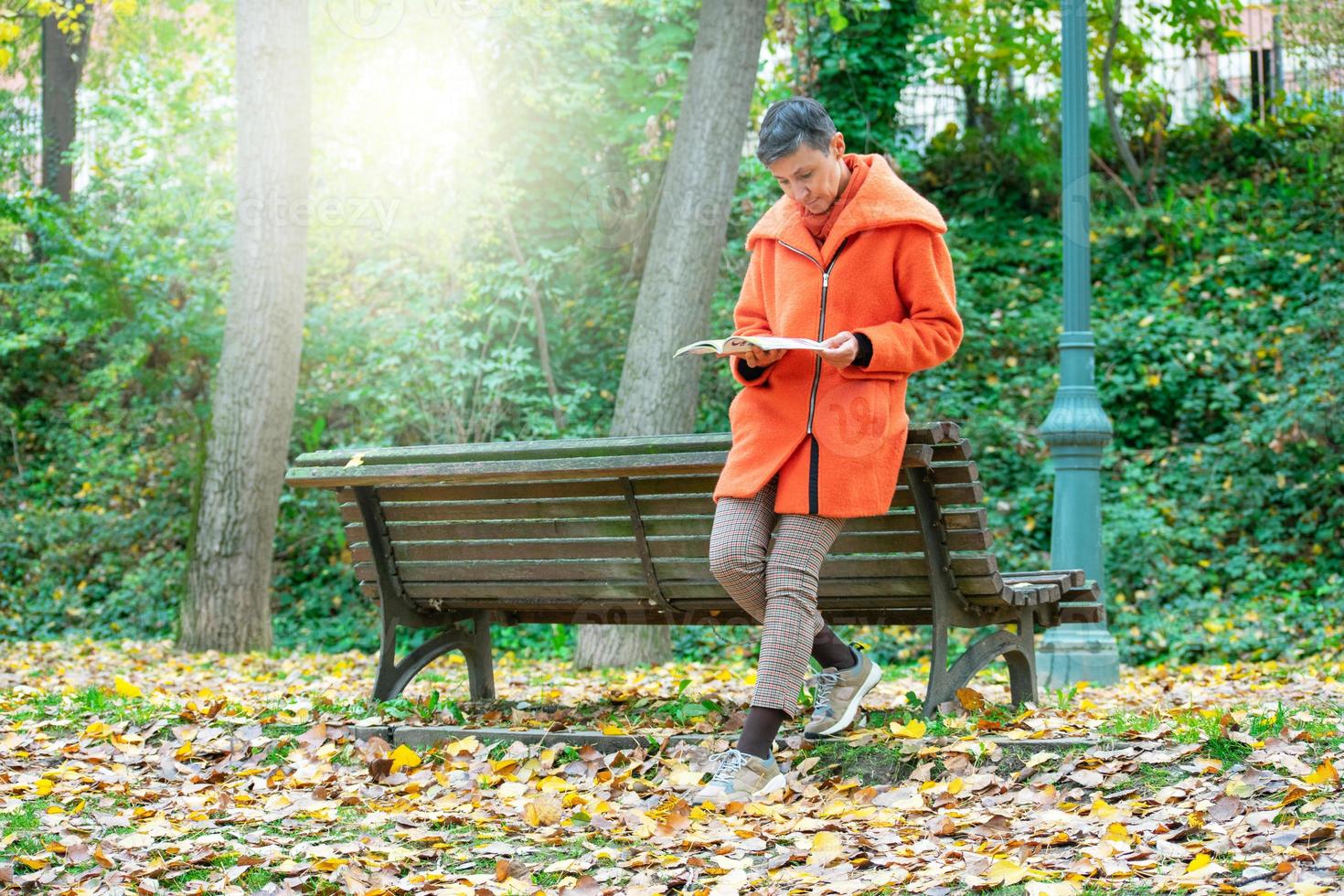 vrouw in oranje jas leest leunend op een bankje in het park foto