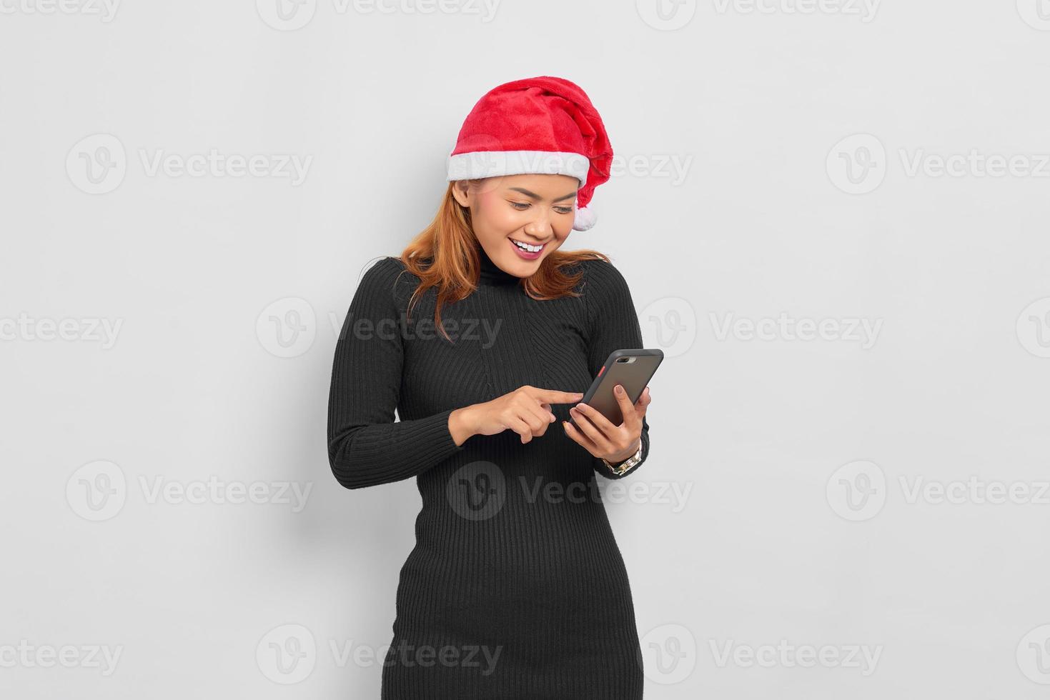 Portret van lachende jonge Aziatische vrouw in kerstman hoed met behulp van mobiele telefoon geïsoleerd op witte achtergrond foto