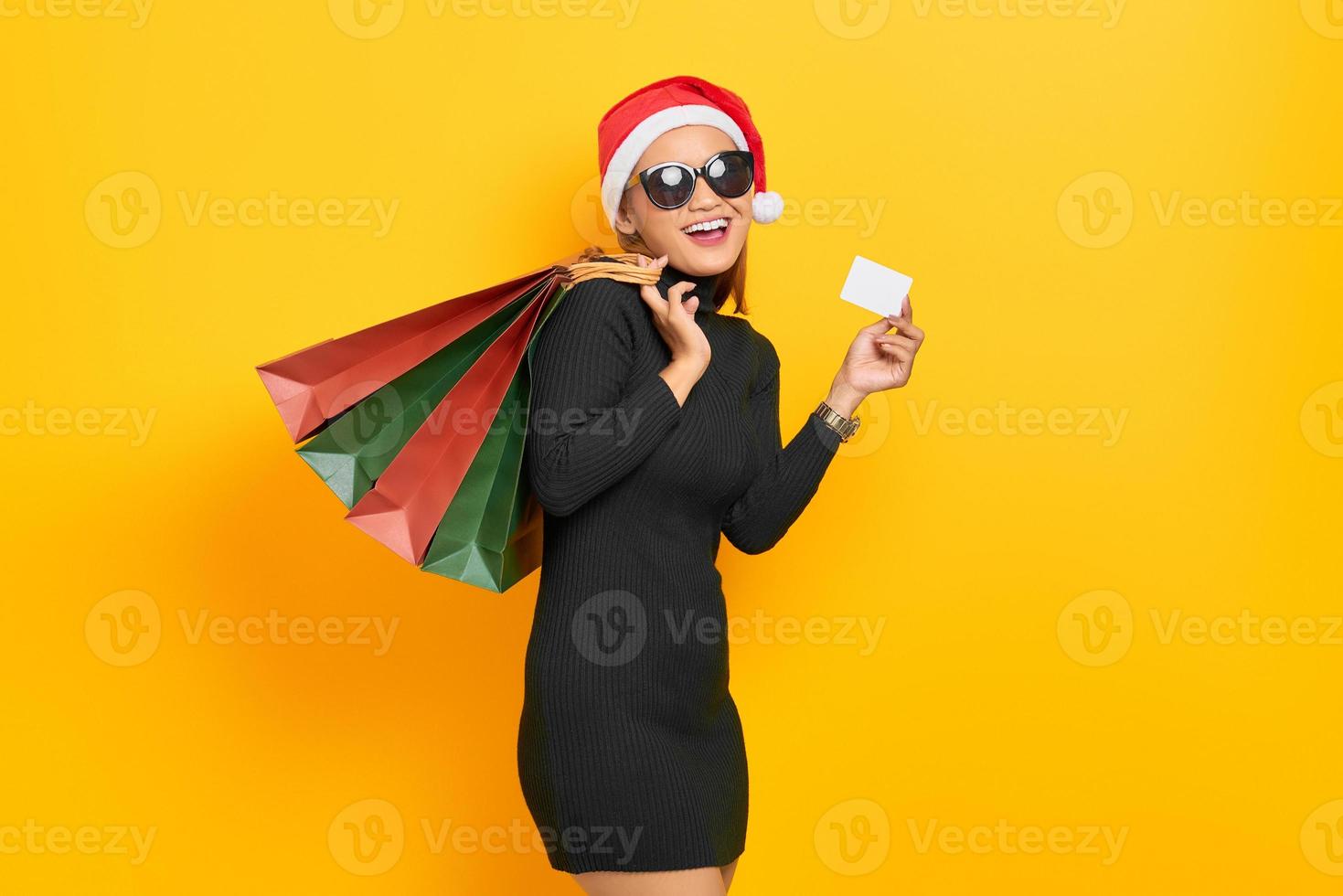 Glimlachende jonge Aziatische vrouw in kerstman hoed en zonnebril met boodschappentassen en plastic kaart geïsoleerd op gele achtergrond foto