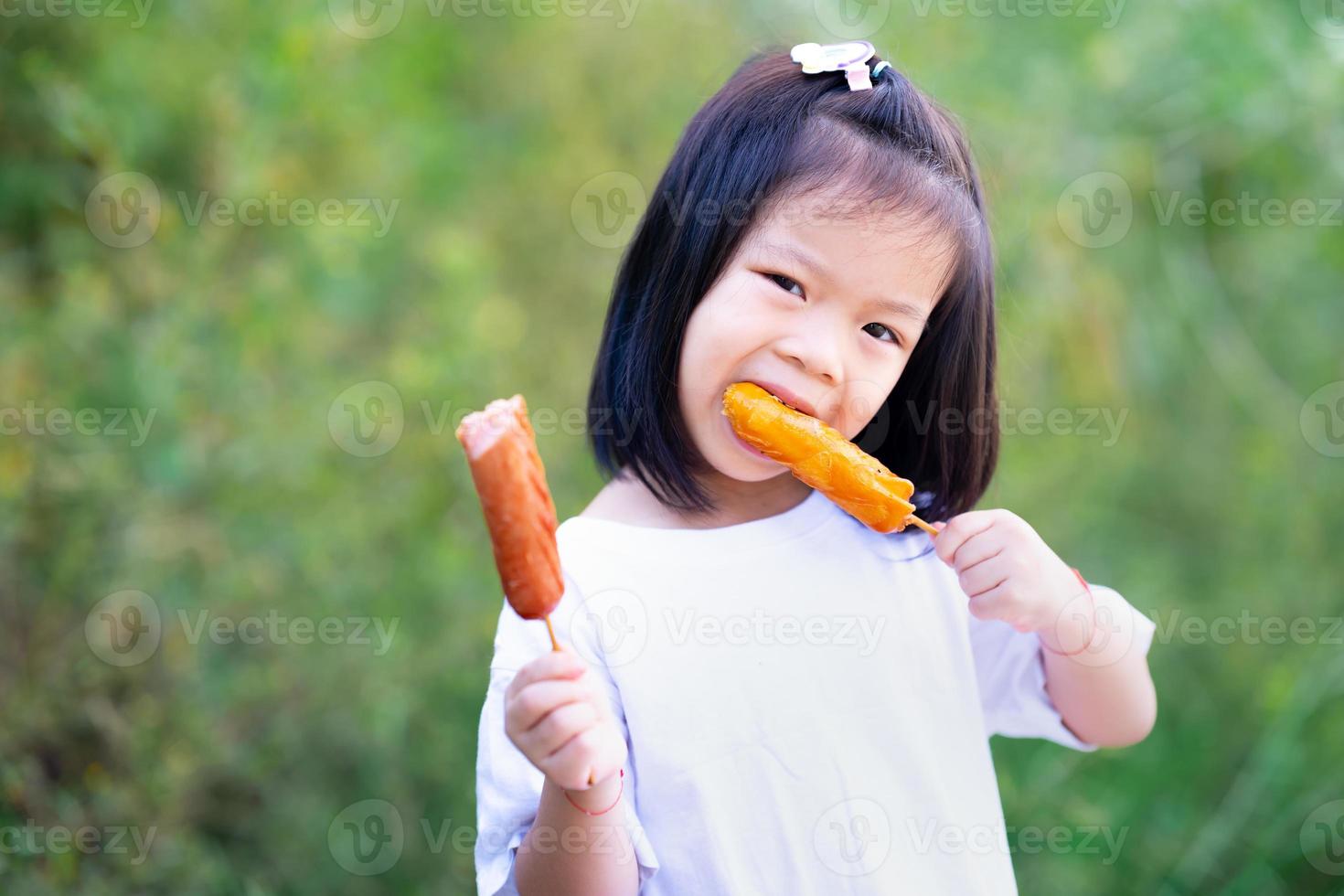 kind houdt twee worstjes in haar handen. schattig meisje at heerlijke spiesmaaltijd. een kind van 4-5 jaar draagt een wit overhemd. kinderen hebben honger. groene natuur achtergrond. lege ruimte om tekst in te voeren. foto