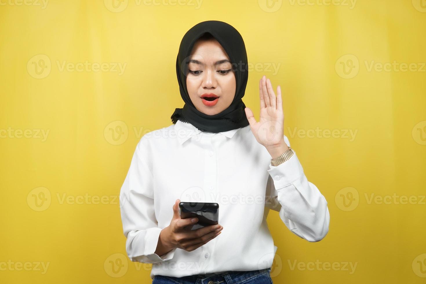 mooie jonge aziatische moslimvrouw geschokt, verrast, met handen met smartphone, kijkend naar smartphone, kijkend naar promo, geïsoleerd op gele achtergrond, reclameconcept foto