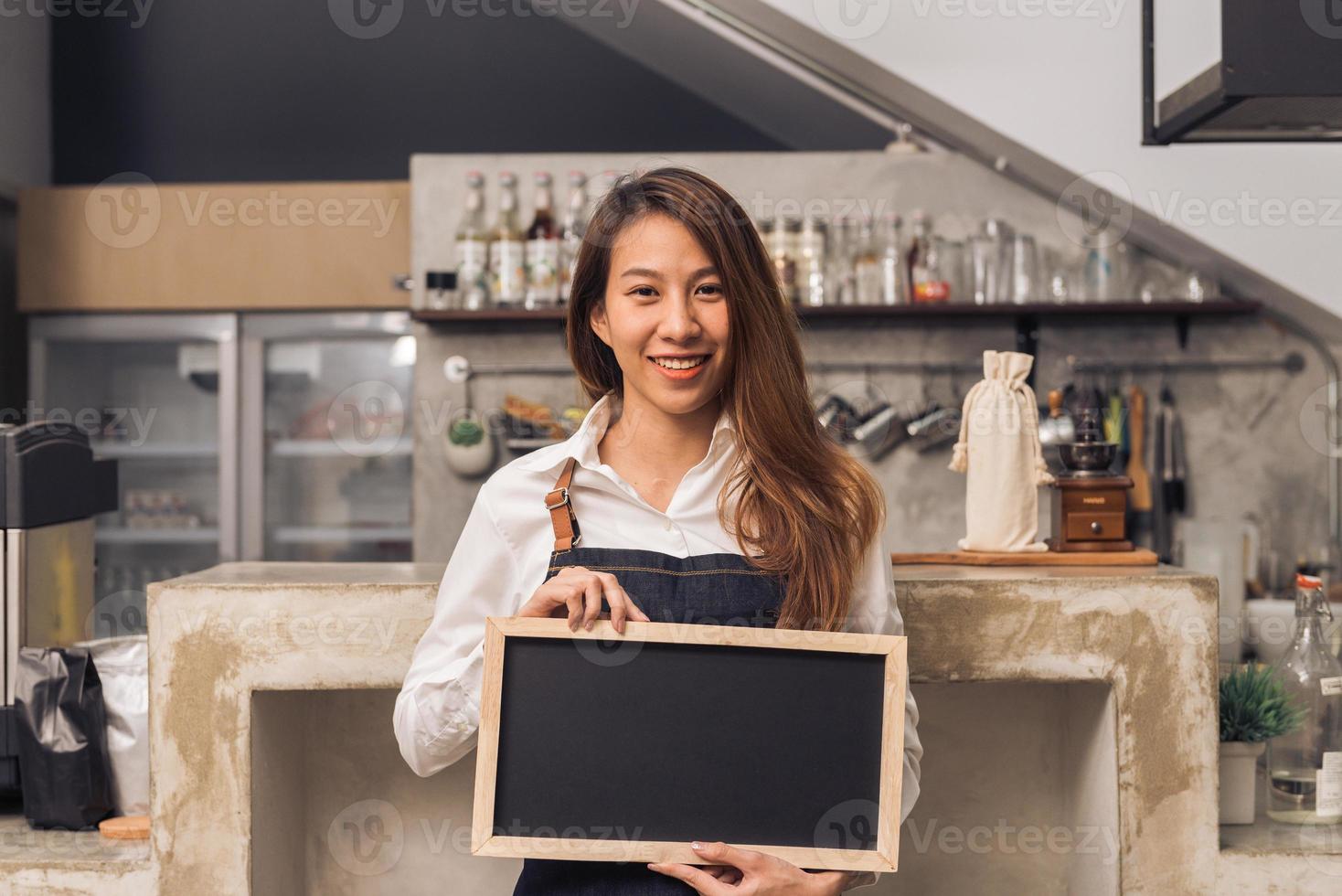 jonge aziatische vrouwelijke barista in denim schort houdt een schoolbord vast met een mooie glimlach in haar eigen coffeeshop verwelkomt haar klant om een nieuw koffiemenu te proberen. jonge vrouwelijke barista en haar kleine koffieshop. foto