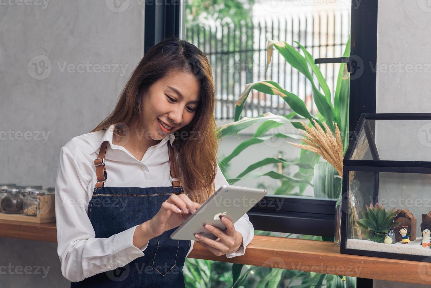 jonge aziatische vrouwelijke barista die elektronische tablet gebruikt in haar eigen coffeeshop terwijl ze een pauze neemt in de warme middag. jonge vrouwelijke barista en haar moderne kleine koffieshop. concept van de voedsel- en drankenindustrie. foto