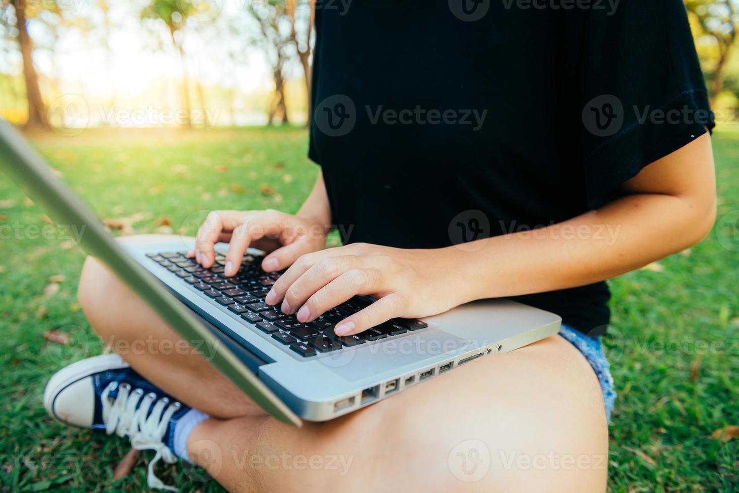 benen van de jonge Aziatische vrouw op het groene gras met geopende laptop. meisje handen op toetsenbord. afstandsonderwijs concept. gelukkig hipster jonge Aziatische vrouw die op laptop in park werkt. student studeert buiten. foto