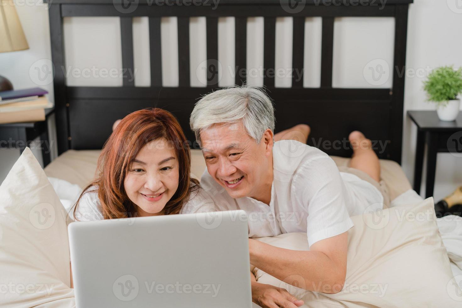 Aziatisch hoger paar dat laptop thuis met behulp van. Aziatische senior chinese grootouders, man en vrouw blij na het wakker worden, kijken naar film liggend op bed in de slaapkamer thuis in het ochtendconcept. foto