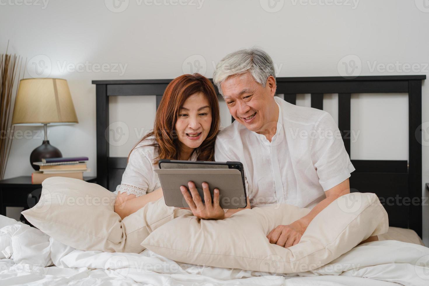 Aziatisch hoger paar dat tablet thuis gebruikt. Aziatische senior chinese grootouders, man en vrouw blij na het wakker worden, kijken naar film liggend op bed in de slaapkamer thuis in het ochtendconcept. foto