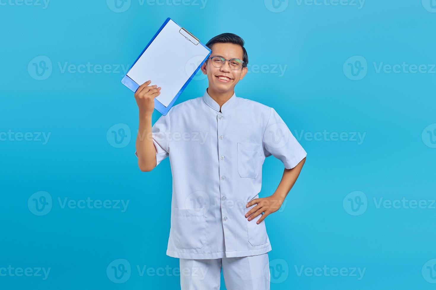 vrolijke jonge verpleger die klembord toont en camera op blauwe achtergrond bekijkt foto