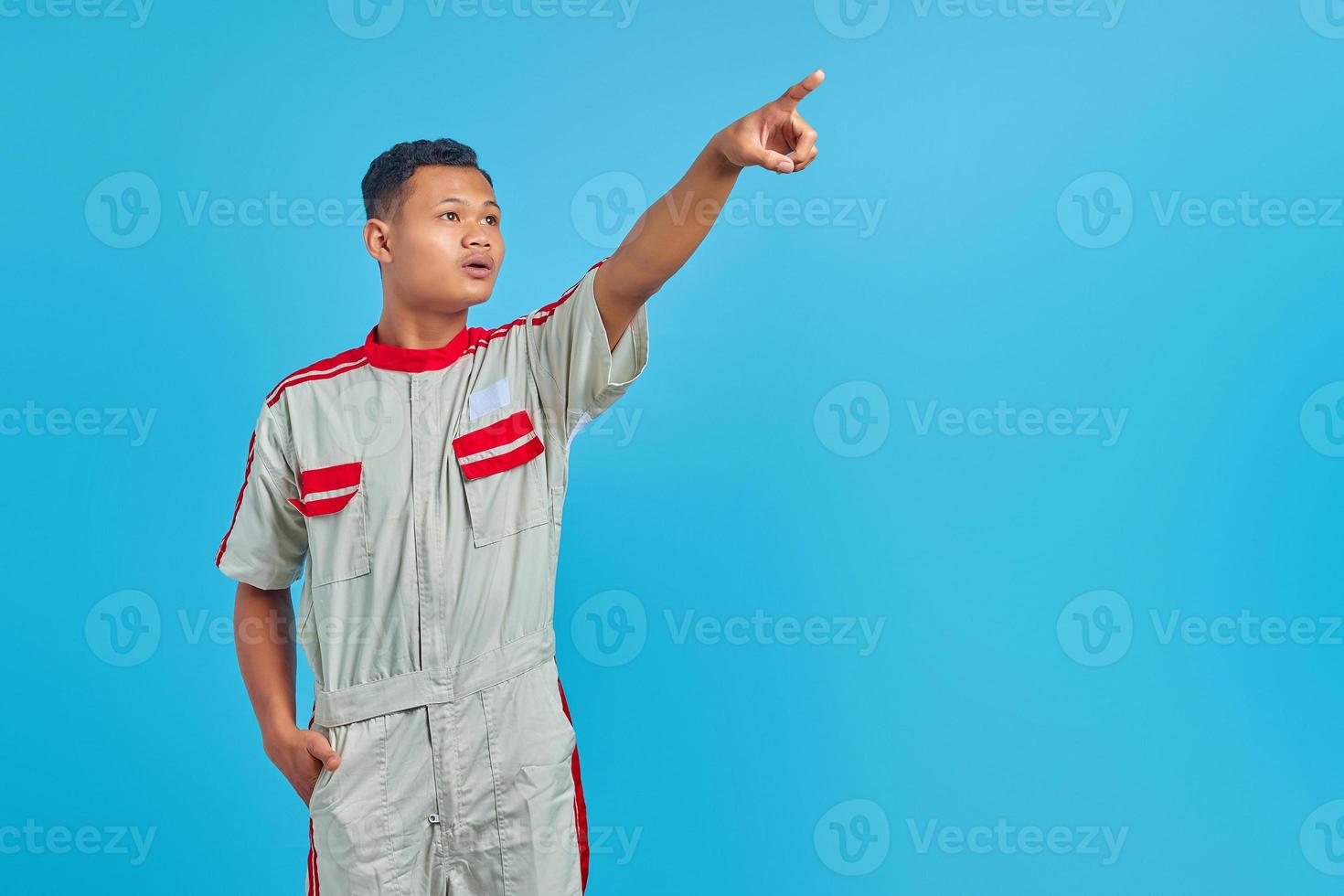 portret van boze jonge Aziatische monteur wijzend op lege ruimte met open vinger en mond over blauwe achtergrond foto