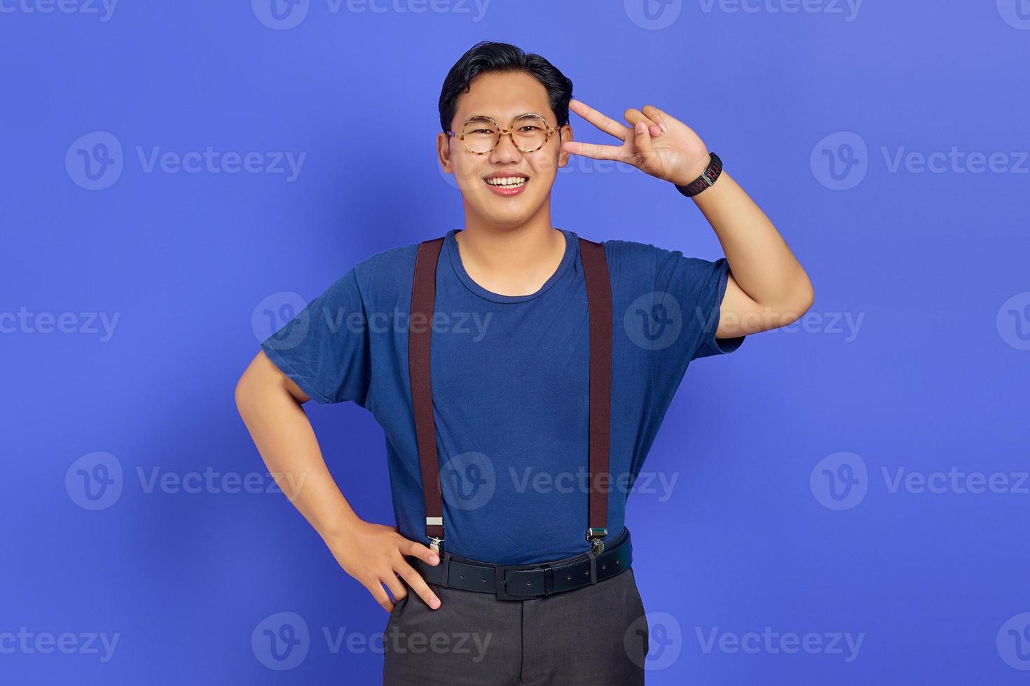 portret van vrolijke aziatische jonge man die vredesteken maakt met vinger over paarse achtergrond foto