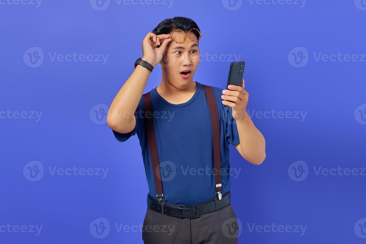 geschokte jonge man die smartphone gebruikt en een bril afzet op een paarse achtergrond foto
