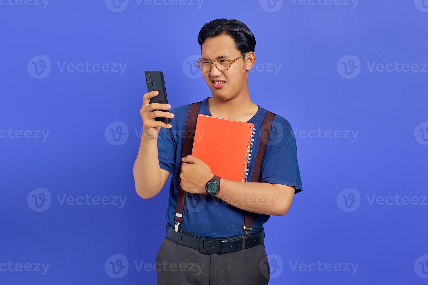 opgewonden jonge knappe student die naar een mobiele telefoon kijkt en een notitieboekje op een paarse achtergrond houdt foto