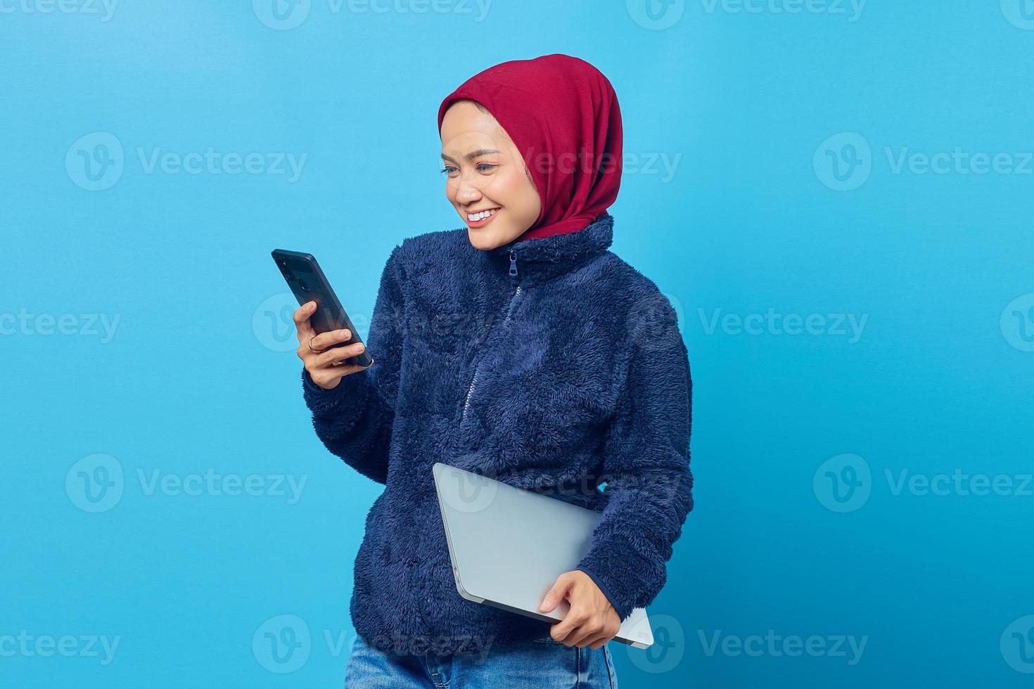 portret van vrolijke jonge aziatische vrouw die smartphone gebruikt terwijl ze de laptop op handen houdt foto