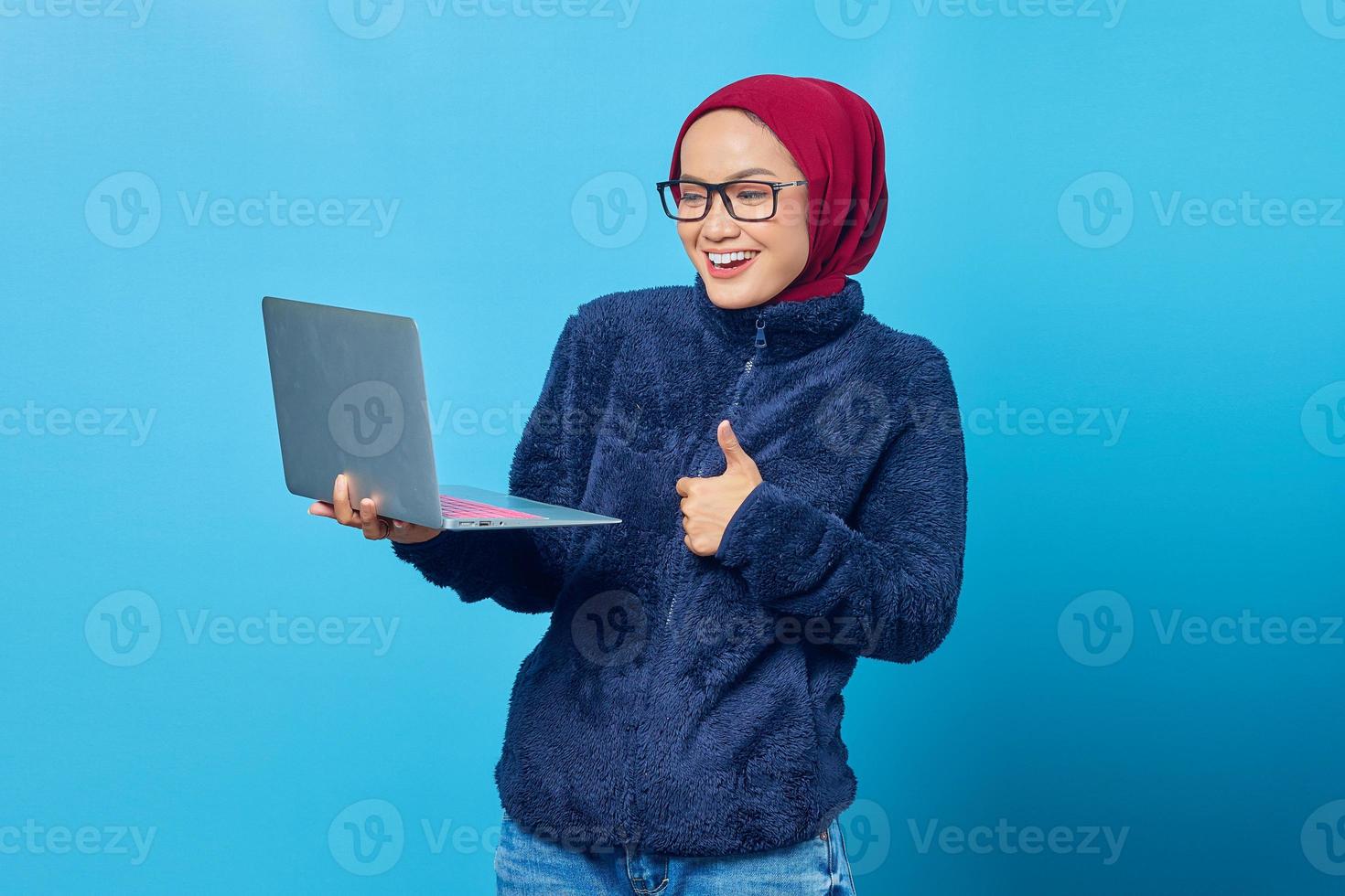 mooie aziatische student die een blauwe jas draagt die laptop draagt en duimen opgeeft met lachende uitdrukking foto