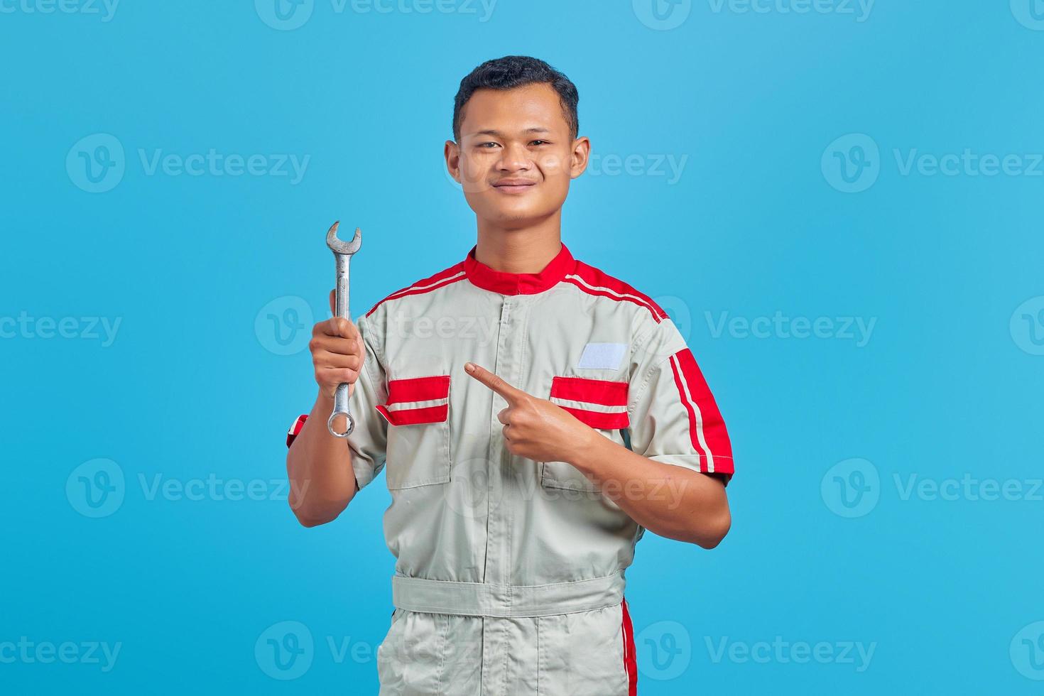 Portret van lachende jonge Aziatische monteur wijzende moersleutel met vinger geïsoleerd op blauwe achtergrond foto