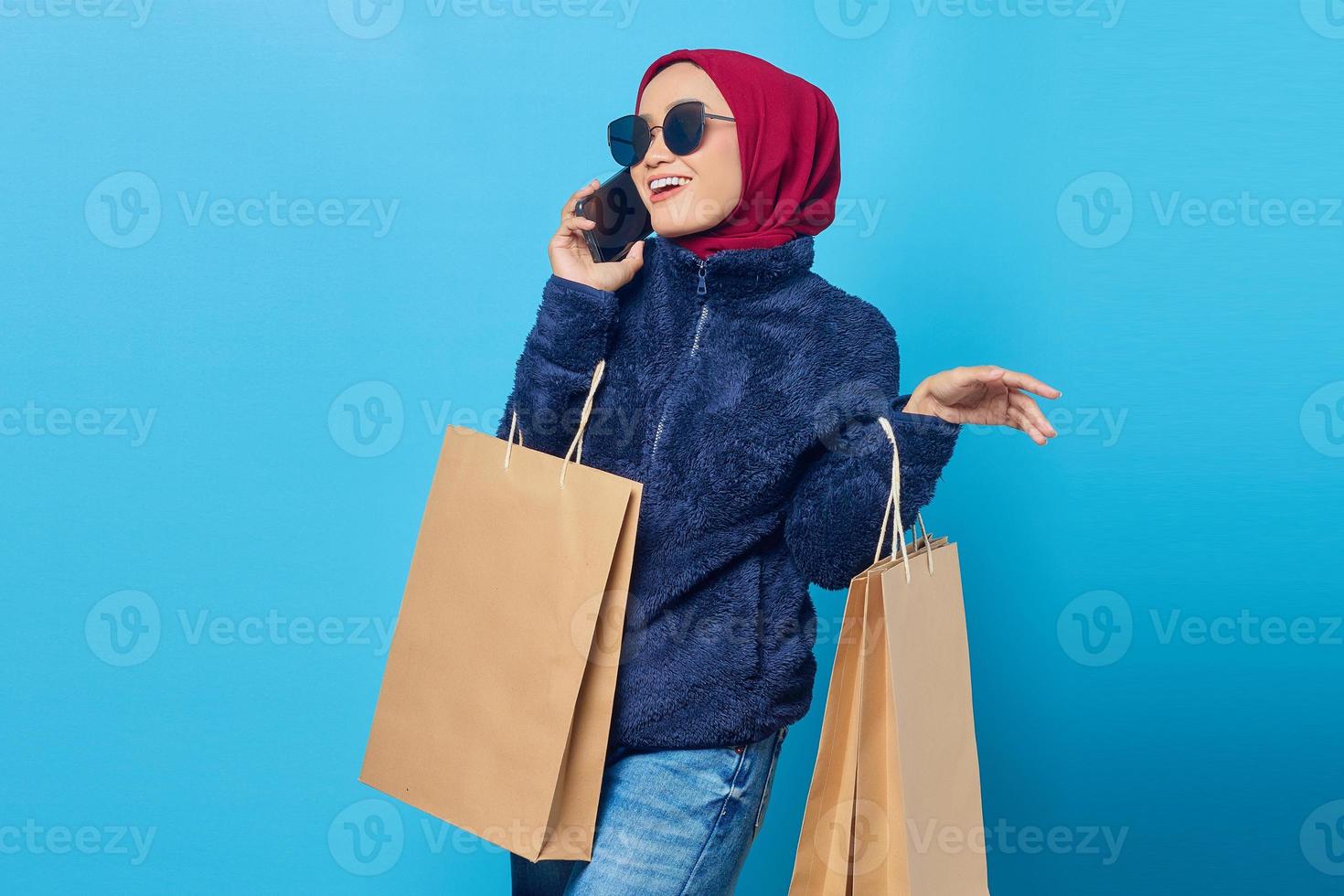 portret van een glimlachende jonge aziatische vrouw die op mobiel praat en boodschappentassen op een blauwe achtergrond vasthoudt foto