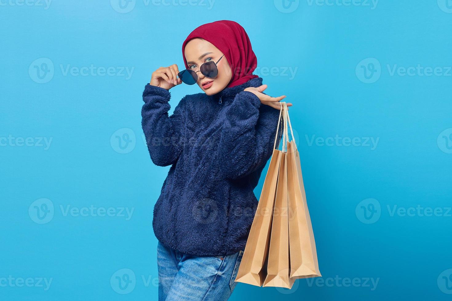 glimlachende jonge aziatische vrouw die boodschappentassen vasthoudt en een bril afzet op een blauwe achtergrond foto