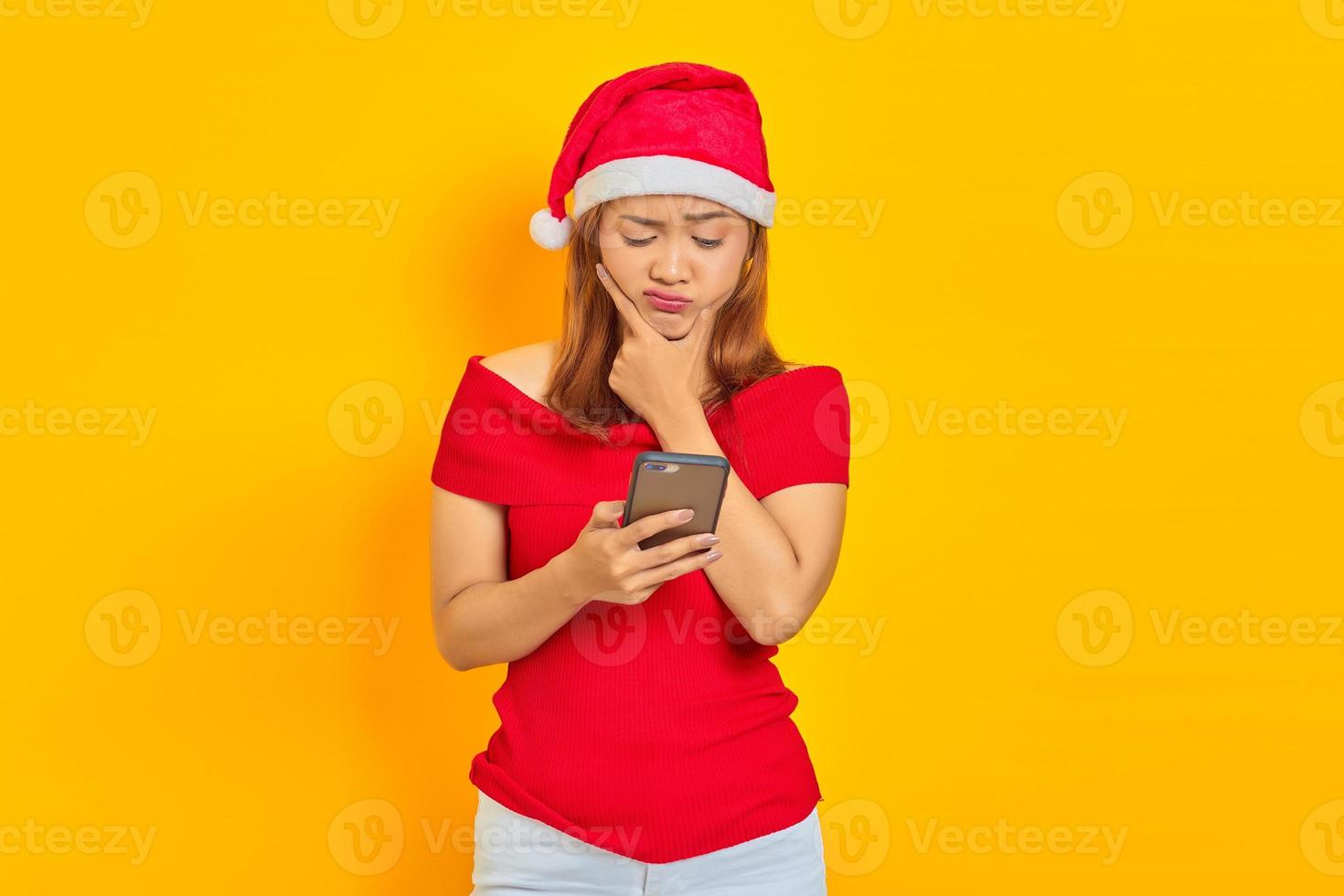 portret van een jonge aziatische vrouw met een kerstmuts die denkt en een bericht verzendt op een smartphone die op een gele achtergrond wordt geïsoleerd foto