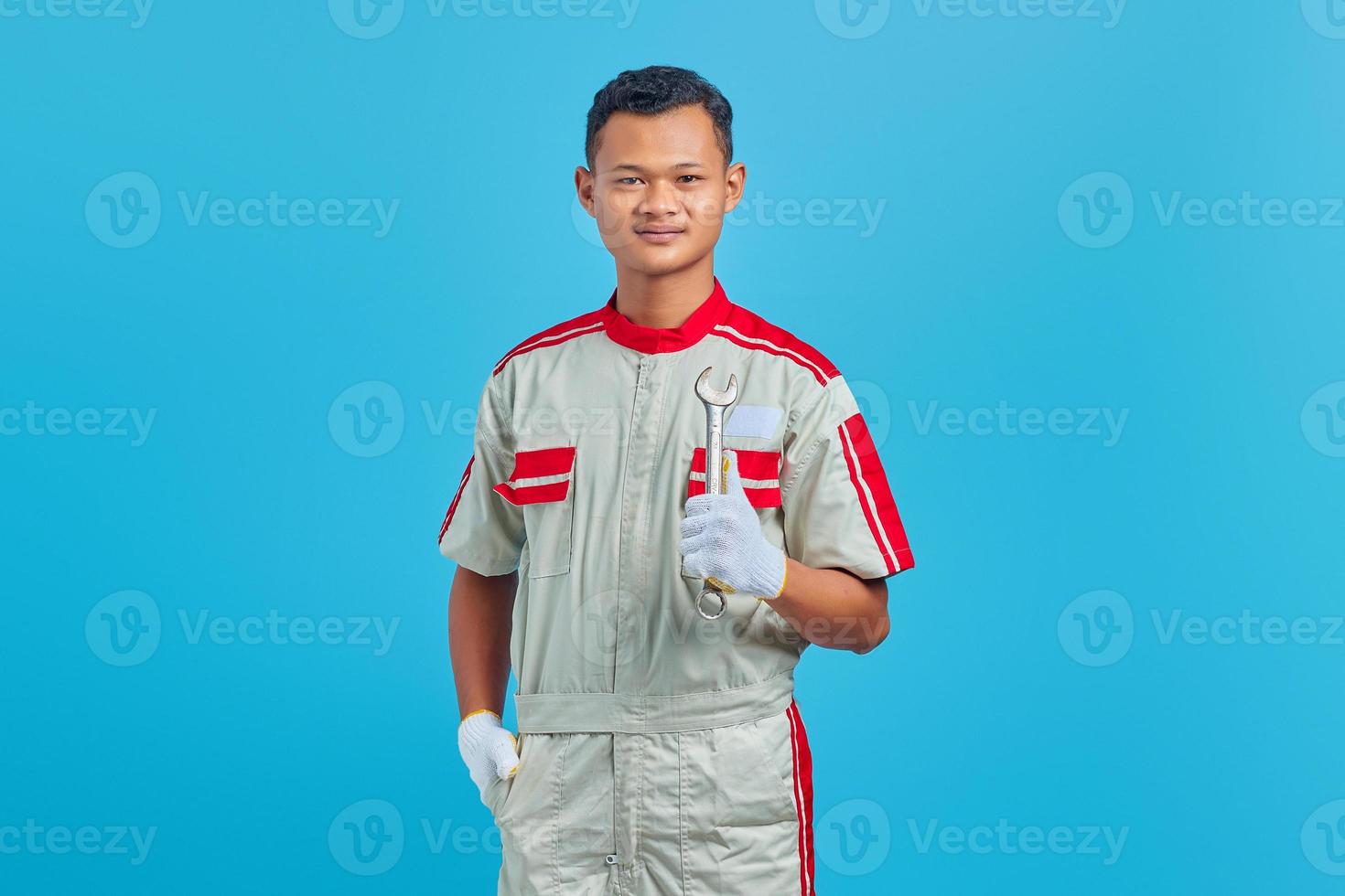 portret van een glimlachende jonge aziatische monteur die een moersleutel in de hand houdt en naar de camera kijkt over een blauwe achtergrond foto