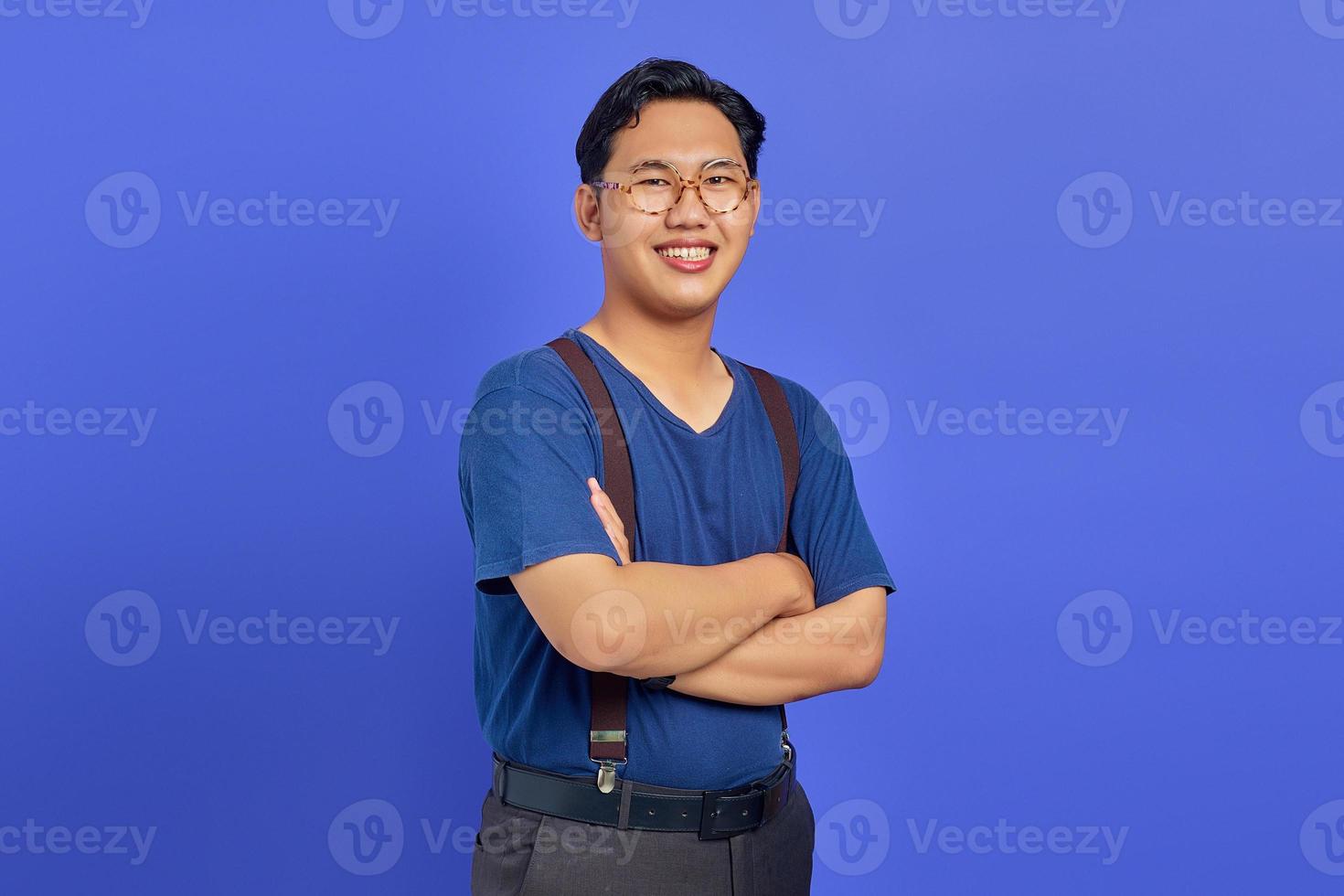 portret van een glimlachende jonge aziatische man die zijn armen kruist en een bril draagt op een paarse achtergrond foto