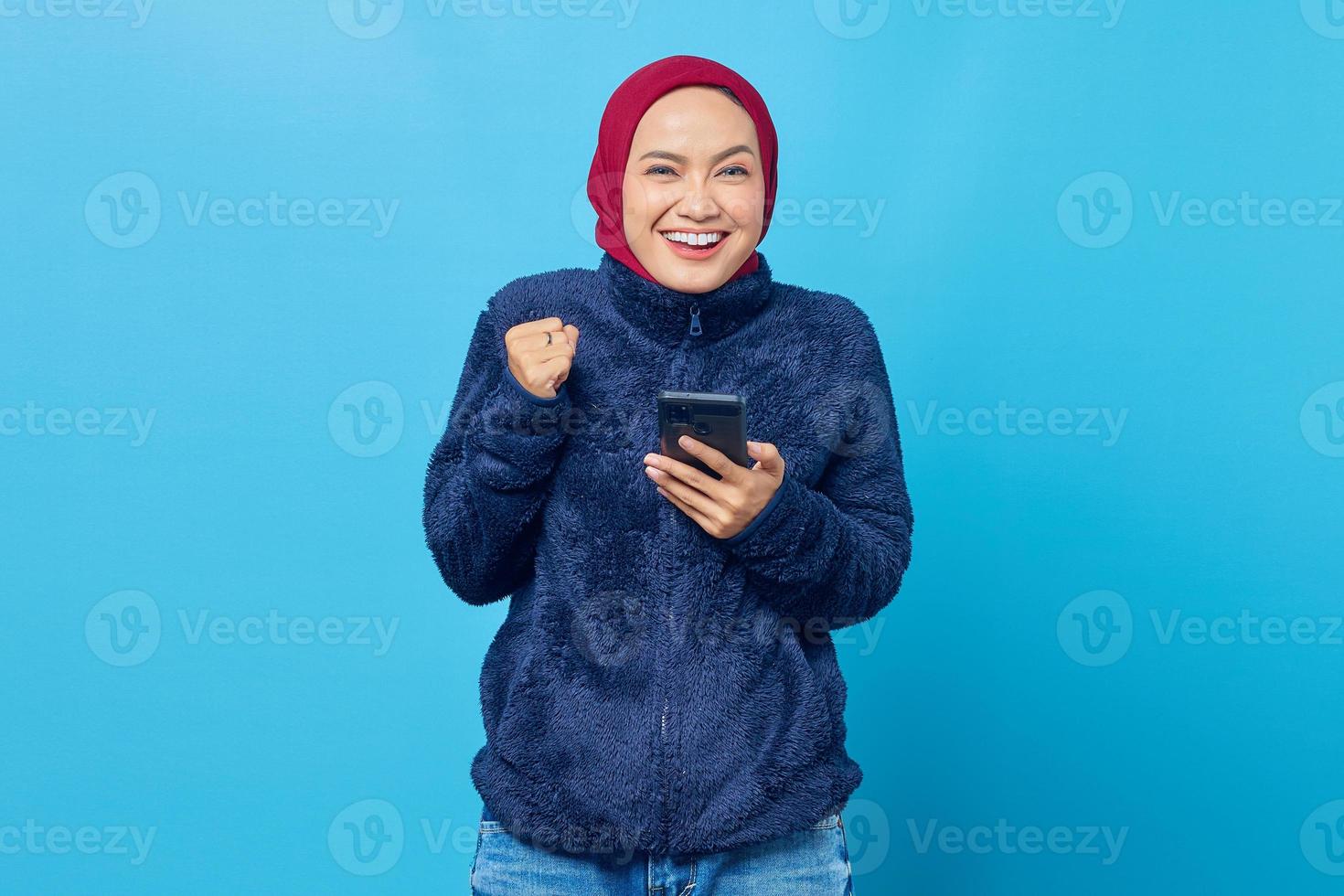 gelukkige jonge aziatische vrouw die mobiele telefoon met succesvol handgebaar op blauwe achtergrond gebruikt foto