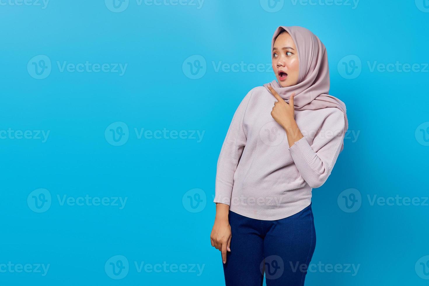 geschokt aziatisch vrouwenportret met vinger wijzend op lege ruimte met open mond over blauwe achtergrond foto