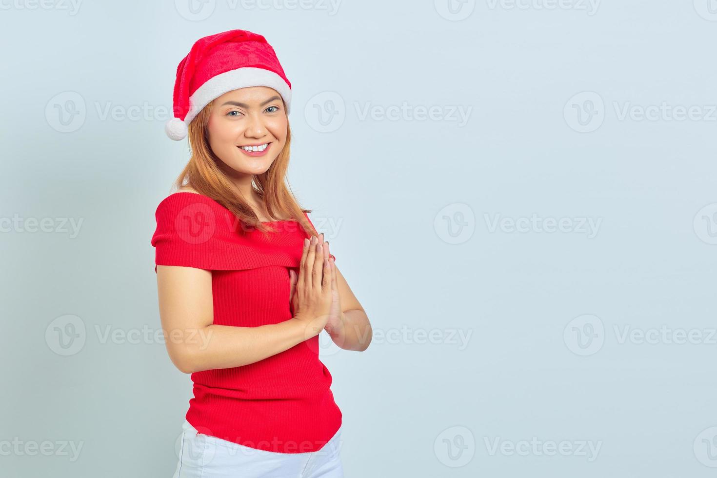 glimlachende jonge aziatische vrouw met kerstmuts die handen vasthoudt en zich optimistisch voelt op een witte achtergrond foto