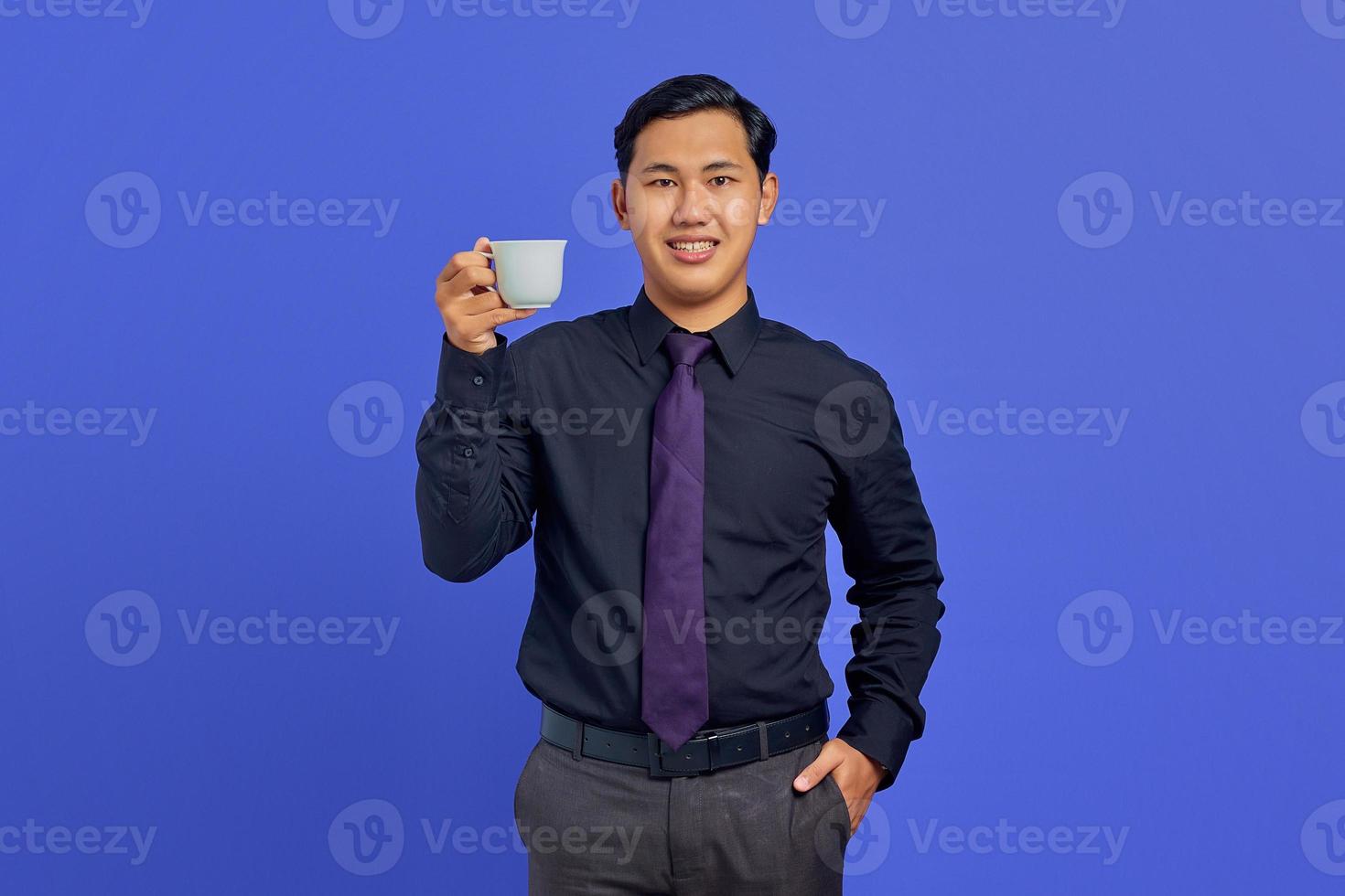 knappe jonge zakenman glimlachend in de camera met handen in zak en mok op paarse achtergrond houden foto