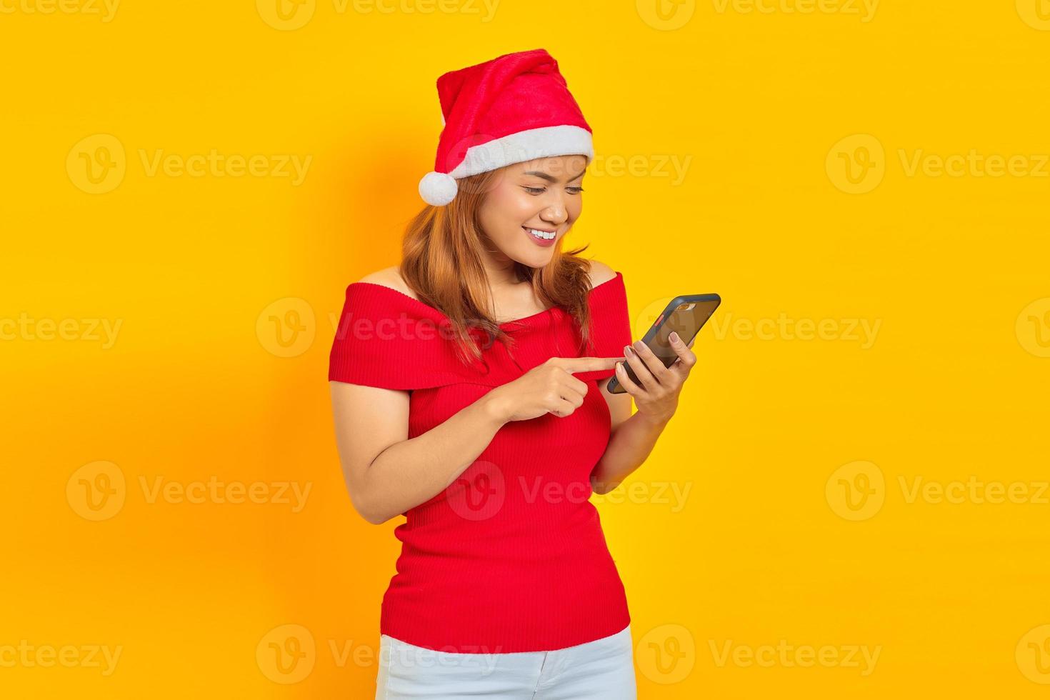 Portret van lachende jonge Aziatische vrouw in kerstman hoed met behulp van mobiele telefoon geïsoleerd op gele achtergrond foto