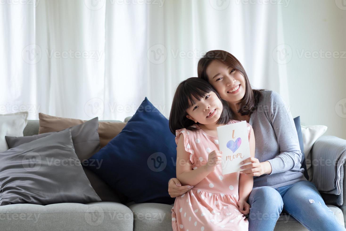 Aziatische moeder knuffelt haar schattige dochter die een handgemaakte wenskaart geeft met het woord 'I love mom' om haar thuis te verrassen foto