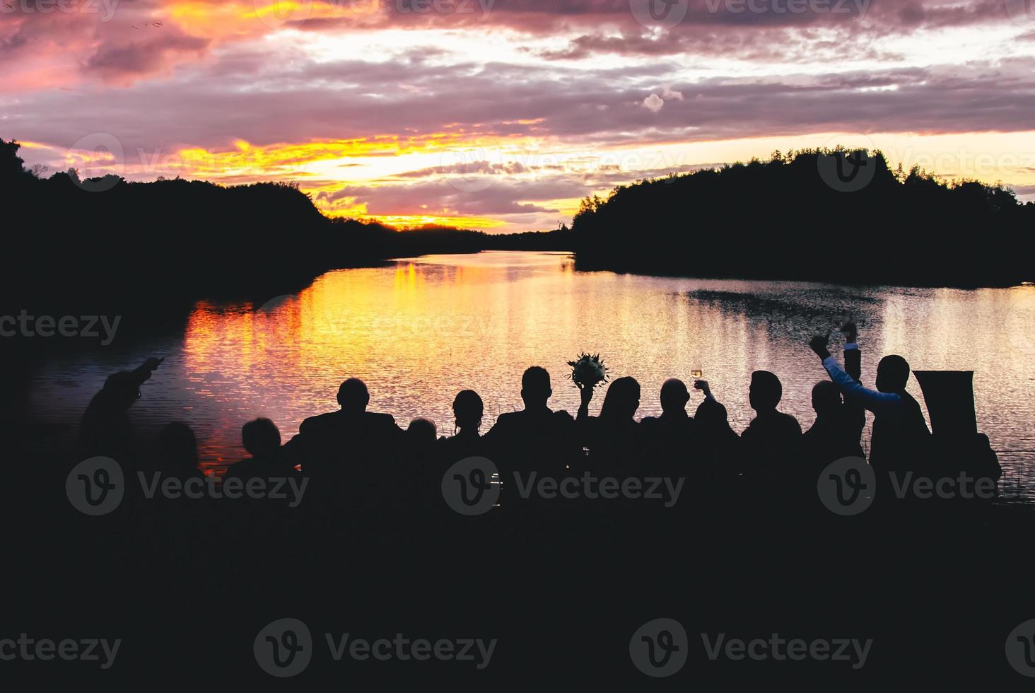 achteraanzicht groep gasten silhouet buiten staan bij meer genieten van zonsondergang op huwelijksfeest in landelijke natuur foto