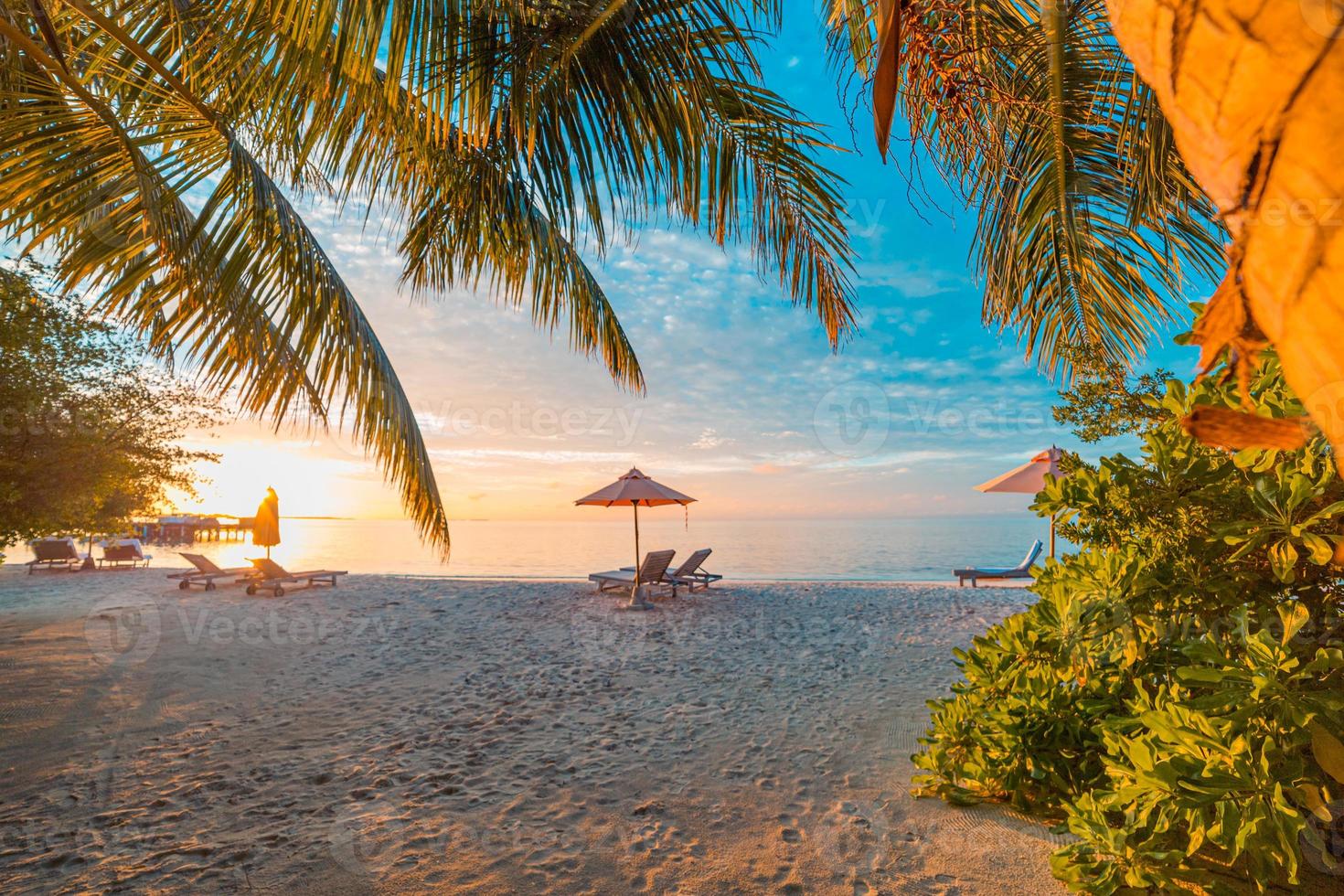 prachtig tropisch zonsonderganglandschap, twee ligbedden, ligstoelen, parasol onder palmboom. wit zand, uitzicht op zee met horizon, kleurrijke schemerhemel, rust en ontspanning. inspirerend strandresorthotel foto