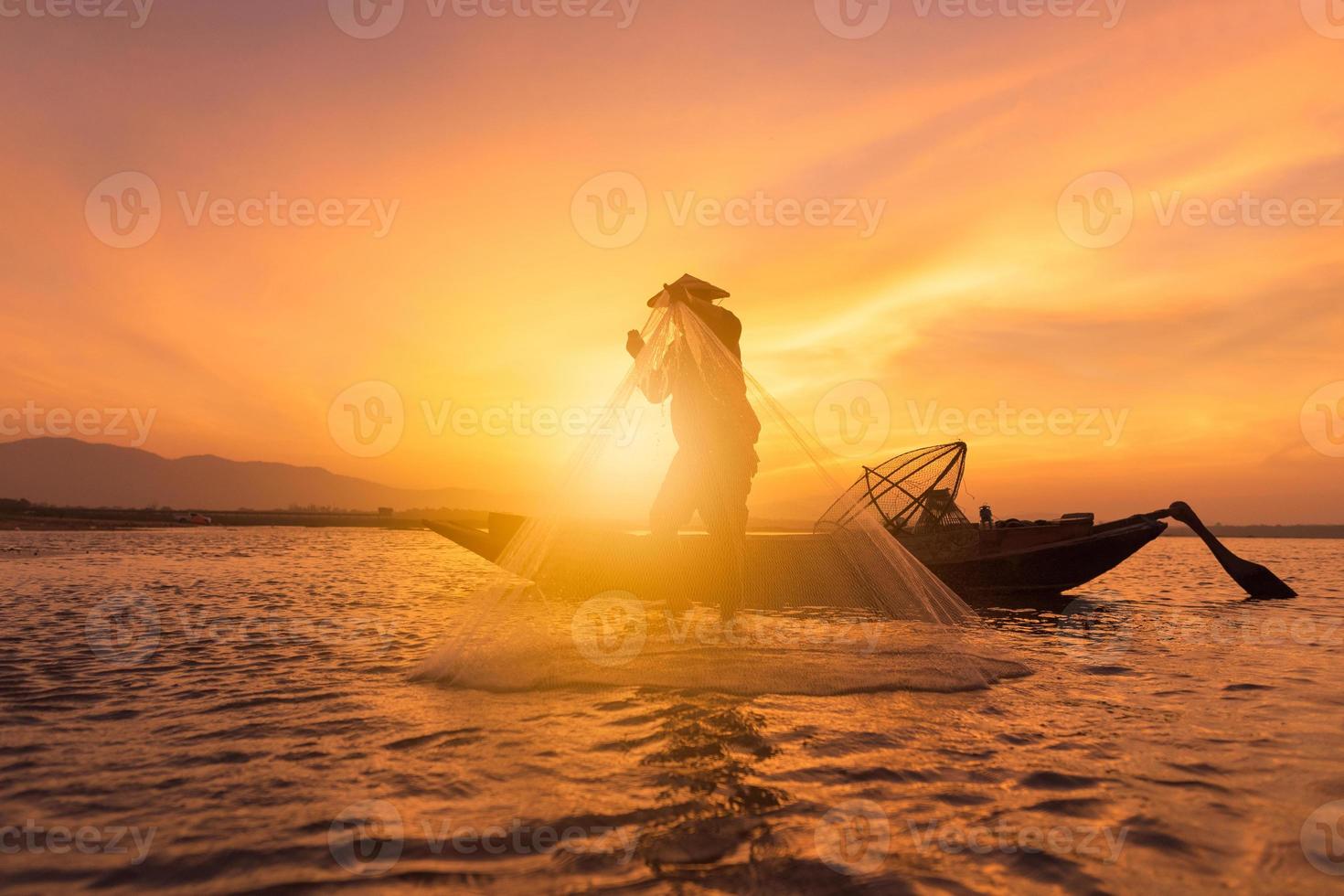 Aziatische visser met houten boot die een net gooit voor het vangen van zoetwatervissen in de natuurrivier in de vroege ochtend tijdens zonsopgang foto